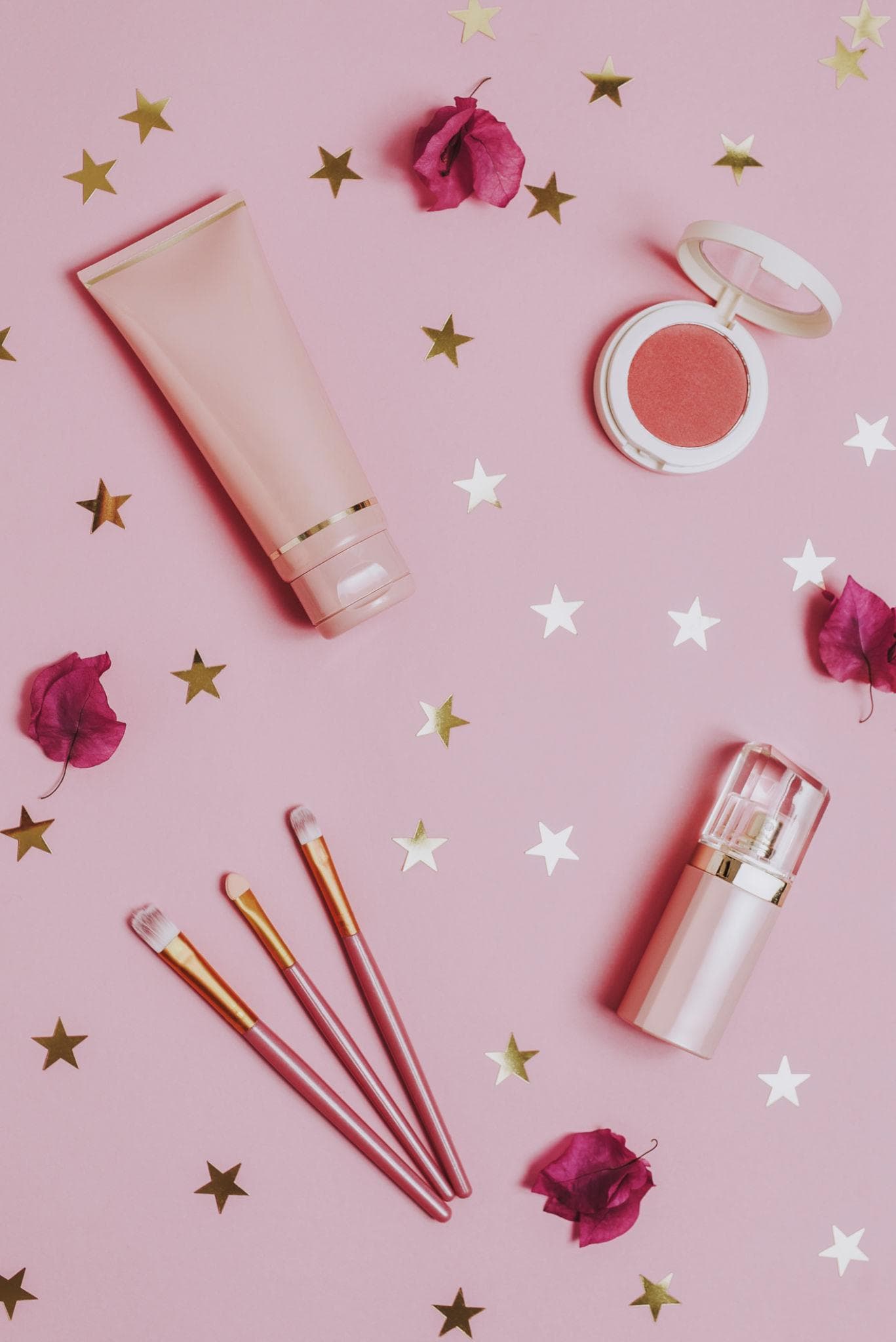 Pink flatlay of lotion, blush, brushes, and a spray bottle with gold star confetti