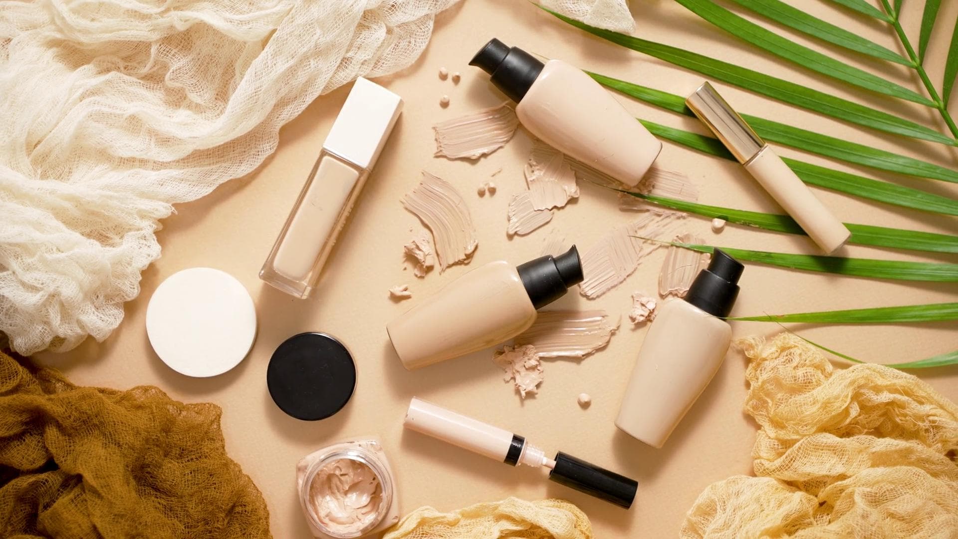 Foundation, concealer, and mascara flatlay on beige with a palm leaf and smudged swatches