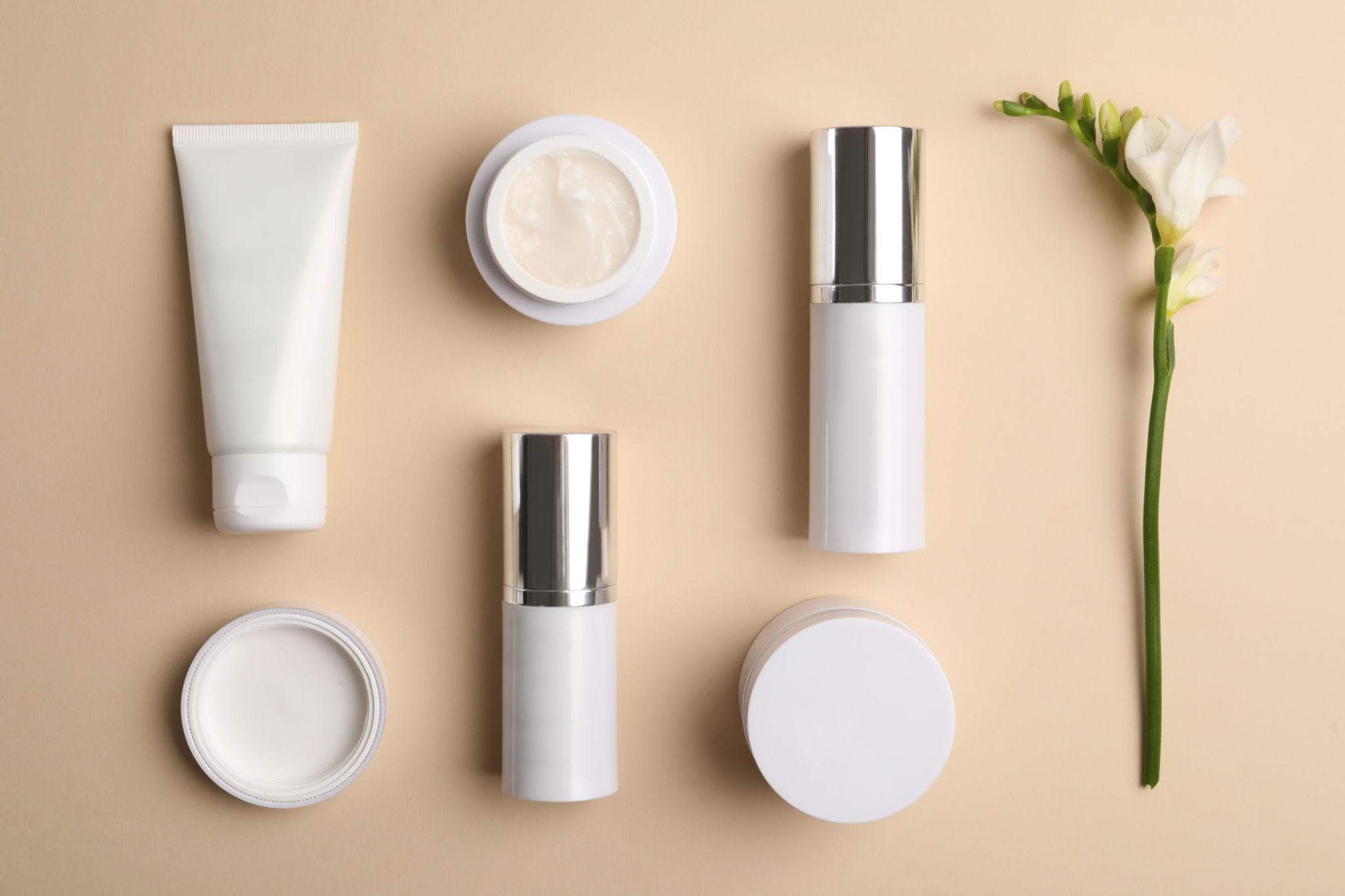 Six white skincare containers - tubes, jars, and bottles - flatlay with a white flower